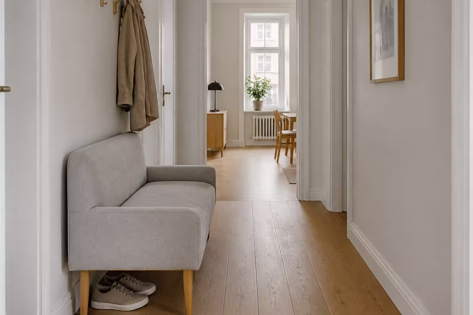 Smal grå tvåsitssoffa längs väggen i en ljus hall med skor under sitsen och krokar ovanför